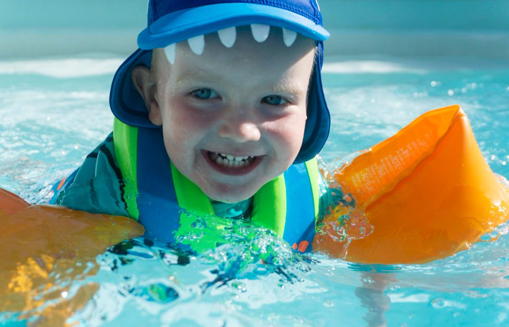 Male toddler with orange Swim Steps swimming arm bands and swimming hat laughing, looking past the camera.